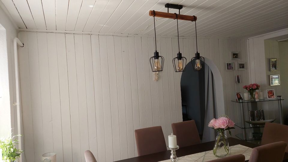 Heller Essbereich mit weißer Holzvertäfelung an Wänden und Decke, einem Esstisch mit Polsterstühlen und einer dreiflammigen Pendelleuchte mit schwarzen Metallkäfig-Schirmen. Im Hintergrund befindet sich ein rundbogiger Durchgang sowie ein Regal mit Deko und Bilderrahmen an der Wand.