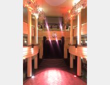 Großer Veranstaltungssaal mit runder Tanzfläche aus Holzparkett, weißen Säulen und zwei umlaufenden Balkonen. Eine Bühne mit schwarzen Vorhängen und eine Disco-Kugel hängen zentral, beleuchtet von bunten Scheinwerfern.