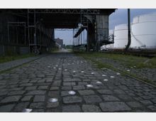 Industriegebiet mit einer Kopfsteinpflasterstraße, die von Metall- und Holzstrukturen sowie teilweise bewachsenen Bereichen gesäumt ist. Im Hintergrund sind weiße Tanks und Gebäude sichtbar, die auf einen Hafen oder ein Lagergelände hinweisen.