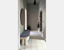 Narrow modern hallway with pale walls and white plank flooring, featuring a cushioned bench and round wall mirror on the left and a black pendant ceiling light overhead. At the far end is a flush door, with coat hooks and a colorful floral wall panel on the right.