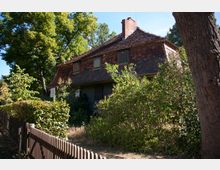 Historisches Haus mit schrägem Ziegeldach und schlichten Fenstern, umgeben von dichtem Grün und Bäumen, mit einem Holzzaun im Vordergrund. Die Umgebung wirkt naturbelassen und von Pflanzen bewachsen.