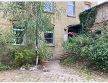 Backsteingebäude mit gelbem Mauerwerk, großen Fenstern und einem kleinen Vorgarten mit Pflastersteinen, Sträuchern, Blumen und Töpfen. Eine kurze Treppe mit Rosensträuchern an der Seite führt zu einer Eingangstür.