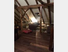 Dachgeschossraum mit sichtbaren Holzträgern und dunklem Holzboden, eingerichtet mit einem kleinen Sitzbereich aus Sesseln und Tisch. Eine Stehlampe spendet warmes Licht, und ein Dachfenster ist im Hintergrund sichtbar.