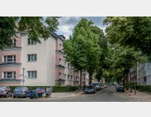 Wohnstraße in einer städtischen Gegend mit mehrstöckigen Wohngebäuden in hellrosa und cremefarbenen Fassaden, flankiert von Bäumen. Am Straßenrand sind parkende Autos, Fahrräder und ein Gehweg zu sehen, der von dichtem Blattwerk überschattet wird.