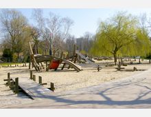Spielplatz mit einem großen Holz-Gerüst in Form eines Schiffs, einer Rutsche und verschiedenen Kletterelementen, umgeben von Sandflächen, Bäumen und einem gepflasterten Fußweg. Im Hintergrund sind weitere Spielgeräte und eine Trauerweide sichtbar.