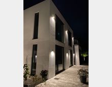 Moderne zweistöckige Wohnhausfassade aus weißem Putz mit großen Fenstern, bei Nacht beleuchtet durch Außenleuchten. Vor dem Haus befindet sich eine gepflasterte Terrasse mit einigen Topfpflanzen und spärlicher Beleuchtung im Hintergrund.