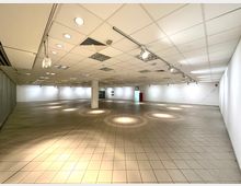 Leerer Innenraum mit weiß gestrichenen Wänden, abgehängter Rasterdecke mit integrierten Leuchten und gefliestem Boden. Ein roter Zugangstürrahmen ist im Hintergrund sichtbar, unterstützt von mehreren Deckenleuchten und Brandschutzdetails.