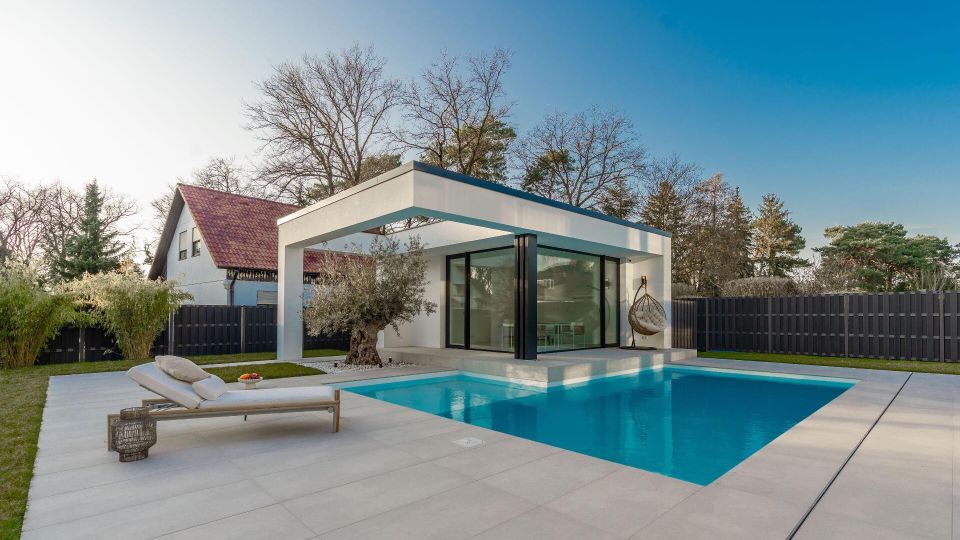 Moderner Poolbereich im Garten mit rechteckigem Swimmingpool, großer heller Terrassenfläche und einer minimalistischen Pavillonstruktur mit Flachdach und bodentiefen Glasfronten. Im Hintergrund stehen ein dunkler Sichtschutzzaun, Bäume und ein Wohnhaus mit Satteldach.