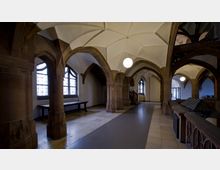 Innenansicht eines historischen Gebäudes mit Kreuzgewölbedecken, Sandsteinsäulen, gotischen Bögen und großen Fenstern mit Gittermustern. Der Raum ist mit Stein- und Holzarbeiten gestaltet und wird durch eine zentrale, hängende Leuchte beleuchtet.