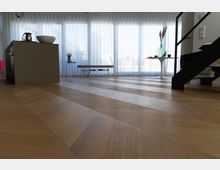 Modernes Wohnrauminterieur mit einem hölzernen Fischgrät-Parkettboden, einer Kücheninsel links, einer Treppe mit schwarzem Geländer rechts und raumhohen Fenstern mit transparenten Vorhängen im Hintergrund. Ein runder Tisch mit Dekoration und einige Möbelstücke ergänzen den Raum.