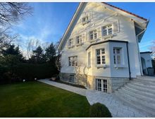 Zweistöckiges Einfamilienhaus in weißem Putz mit roten Dachziegeln und weißen Fensterläden, umgeben von einem gepflegten Garten mit Rasen, Hecken und Baumschnitt; eine Steintreppe führt zur Haustür. Im Vordergrund eine gepflasterte Fläche und eine niedrige Natursteinfassade am Sockel des Gebäudes.