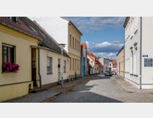 Gepflasterte Straße in einer Wohngegend mit niedrigen Häusern in verschiedenen Pastellfarben, einige mit Blumen an den Fenstern. Am Straßenrand sind geparkte Autos zu sehen, und im Hintergrund erstreckt sich der Straßenzug unter einem bewölkten Himmel.