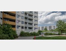 Mehrstöckige Wohngebäude mit Balkonen und weißen Fassaden, umgeben von grünen Büschen und Bäumen. Ein Gehweg führt durch eine mit Rasen und vereinzelten Bäumen gestaltete Umgebung, unter einem teilweise bewölkten Himmel.