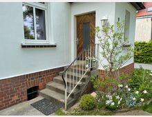 Eingangsbereich eines Hauses mit weißer Fassade und rotem Klinkersockel. Eine Betontreppe mit Metallgeländer führt zu einer Holztür, daneben ein kleiner Garten mit Blumen, Sträuchern und Rasen.