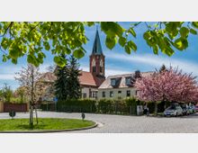 Eine kleine Kirche mit einem Spitzdach und Uhrenturm steht inmitten eines Dorfes, umgeben von Wohnhäusern, blühenden Kirschbäumen und einem gepflasterten Platz. Im Vordergrund sind grüne Blätter und ein runder, bepflanzter Verkehrsbereich zu sehen.