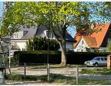 Wohngebiet mit Einfamilienhäusern, teils mit dunklen und roten Dächern, umgeben von Bäumen und Hecken. Im Vordergrund ein Metallzaun, ein sandiger Weg und eine Straße mit gepflastertem Bürgersteig, daneben steht ein parkendes Auto.