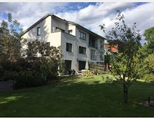 Mehrstöckiges Wohnhaus in moderner Bauweise mit hellgrauer Fassade, großen Fenstern und Balkonen, umgeben von einem Garten mit Rasen, Bäumen und Sträuchern. Im Vordergrund befindet sich ein Essbereich mit Sonnenschirm.