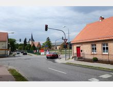 Straßenkreuzung in einem Dorf mit einem roten Backsteinhaus und roten Dachziegeln rechts im Vordergrund. Im Hintergrund sind eine Kirche mit Turmspitze sowie weitere niedrigere Gebäude und Bäume zu sehen.