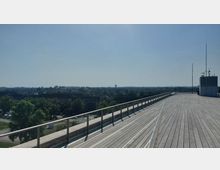 Weitläufige Dachterrasse mit Holzplankenboden und Metallgeländer, die auf eine urbane Landschaft mit Gebäuden, Bäumen und einem fernen Hochhaus blickt; im Hintergrund ein klarer Himmel.