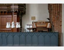 Innenraum einer Kirche mit einer Holzorgel im Hintergrund, flankiert von roten Backsteinmauern und einer hölzernen Kanzel mit geschnitzten Details. Im Vordergrund eine dunkelgraue Balustrade mit gotischen Verzierungen sowie ein moderner Kronleuchter.