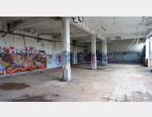 Ein leerstehender, heruntergekommener Innenraum eines Gebäudes mit gefliesten Böden, Betonpfeilern und Wänden, die mit bunten Graffitis bedeckt sind. Die Decke weist sichtbare Schäden auf, und vereinzelte Rohre ragen hervor.