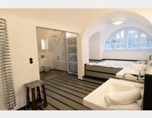 Modernes Badezimmer mit gewölbter Decke, gestreiftem Fliesenboden und großem Rundbogenfenster. Rechts stehen eine eingelassene Eckbadewanne und ein Doppelwaschbecken; links befindet sich eine verglaste Dusche mit Handtuchheizkörper.