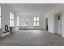 Leerstehender, heller Raum mit weißen Wänden, großen Sprossenfenstern, Betonboden und sichtbaren Heizungsrohren. Rechts befindet sich eine Tür mit goldenem Griff, und an der linken Wand ist eine graue Schlauchskulptur angebracht.