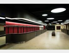 Moderner Garderobenbereich mit dunklen Tresen, rot hinterlegten Wandpaneelen und einer langen Reihe an Haken. Der Raum hat eine helle Steinbodenfläche, eine dunkle Decke mit großen weißen Leuchtkreisen und einen rechteckigen Sitzblock in der Mitte.