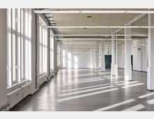 Großer, leerer Innenraum mit hohen Decken, weißen Wänden und vielen bodentiefen Fenstern auf der linken Seite, die viel Tageslicht hereinlassen. Die Decke verfügt über sichtbare Kabeltrassen und schmale, rechteckige Leuchten, während der Boden aus glattem Beton besteht.