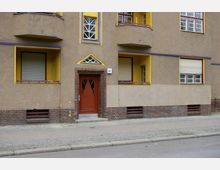 Fassade eines Wohngebäudes mit gelb abgesetzten Fenstern und Balkonen. Die Haustür ist rot, von braunen Ziegelsteinen umrahmt und dreieckige Fenster sind darüber in die Wand integriert; der Gehweg ist aus Pflastersteinen.