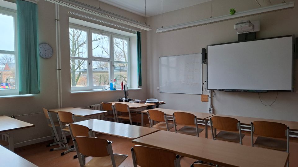 Leerer Klassenraum mit Reihen aus Holztischen und -stühlen, großen Fenstern mit grünen Vorhängen und einer Wanduhr. Vorn befinden sich eine Tafel bzw. ein interaktives Whiteboard mit Deckenprojektor sowie ein Lehrerpult.