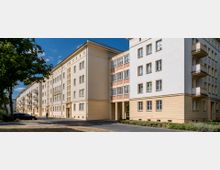 Mehrstöckiges Wohngebäude im Stil der Nachkriegsarchitektur mit hellgelber und weißer Fassade, regelmäßigen Fensterreihen und Balkonen an der Seite. Im Vordergrund sind ein Gehweg, eine Rasenfläche und einige Bäume sichtbar, bei klarem Himmel und Tageslicht.