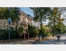 Straßenszene in einem Wohngebiet mit gepflastertem Gehweg, alten Villen mit verzierten Fassaden im Hintergrund und einer Radfahrerin im Vordergrund. Die Straße ist gesäumt von Bäumen und Zäunen mit üppiger Begrünung.