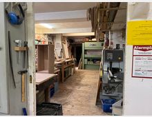 Eine Tischlerwerkstatt mit Holzboden, Werkbänken, hölzernen Regalen und Werkzeugen. Im Vordergrund hängen Handwerkzeuge, während Maschinen und Holzarbeiten im Hintergrund sichtbar sind.