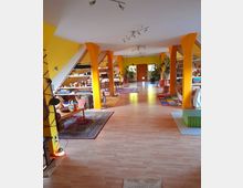 Ein großer, heller Raum mit gelb gestrichenen Wänden und sichtbaren Holzbalken, der als gemütlicher Wohn- oder Arbeitsbereich genutzt wird. Der Raum ist mit Bücherregalen, Teppichen, Pflanzen und verschiedenen Sitzmöglichkeiten eingerichtet, und es gibt breite Fenster sowie Holzböden.