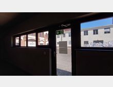 Blick von innen durch eine Glas-Notausgangstür mit Fluchtweg-Schild nach außen, wo ein Hof mit Gebäuden sichtbar ist; die linke Fassade zeigt Ziegelstein-Elemente, während die rechte Wand Graffiti aufweist.