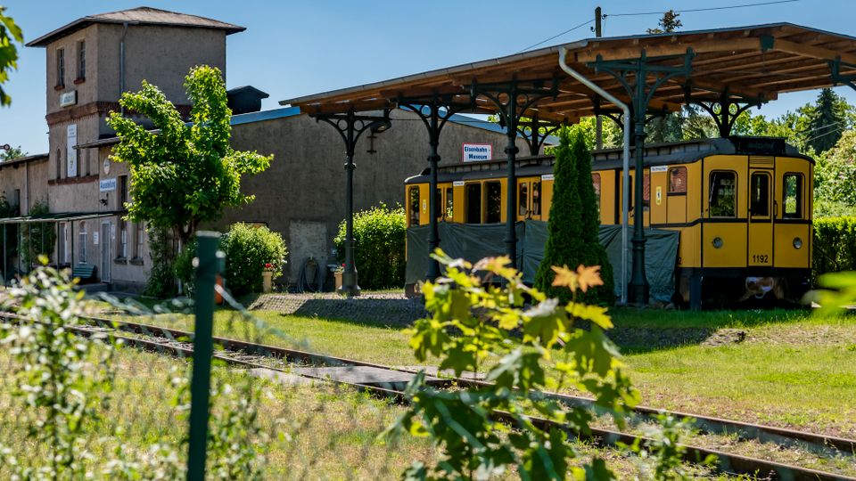 Kleiner, ländlicher Bahnhof mit Gleisen im Vordergrund, einem älteren Stationsgebäude und Signaltechnik. Rechts steht unter einem überdachten Bahnsteig mit gusseisernen Stützen ein gelber historischer Triebwagen, umgeben von Grünflächen und Bäumen.