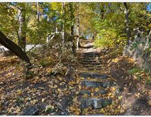 Ein steiler, steinerner Treppenweg führt durch einen herbstlichen Wald mit gelben und braunen Laubblättern am Boden. Links steht ein Baum, während rechts ein Maschendrahtzaun den Weg begrenzt.