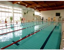 Innenansicht eines Schwimmbads mit einem großen rechteckigen Becken, das durch Schwimmbahnen mit rot-weißen Leinen unterteilt ist. Hohe Fenster sorgen für viel Tageslicht, und die Decke besteht aus sichtbaren Holzträgern; an den Seitenwänden befinden sich Pflanzen und Sitzgelegenheiten.