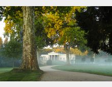 Eine weiße, klassizistische Villa mit Säulenfassade steht im Hintergrund, umgeben von einem gepflegten Park mit großen, dicht belaubten Bäumen. Ein geschwungener Kiesweg führt durch die grüne Wiese, während im Vordergrund ein mächtiger Baumstamm zu sehen ist; leichter Nebel schwebt über der Landschaft.