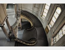 Große, geschwungene Treppe in einem historischen Gebäude mit verzierten Geländern und hohen, mit Buntglasfenstern ausgestatteten Wänden, die weiches Licht in den Raum lassen. Die Architekturelemente umfassen dekorative Säulen und eine helle Wandverkleidung.