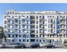 Mehrstöckiges modernes Wohngebäude mit weißer Fassade, regelmäßigen Balkonen und großen Fenstern; davor verläuft eine Straße mit parkenden und vorbeifahrenden Autos sowie einer Haltestelle mit einem überdachten Wartebereich.