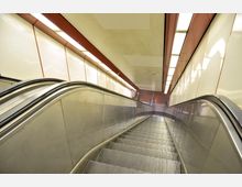 Rolltreppe in einer U-Bahn-Station mit beleuchteten Wänden aus glatten weißen Paneelen und roten Akzenten an der Decke. Die Rolltreppe führt nach unten, und der Bodenbereich ist mit braunen Fliesen gestaltet.