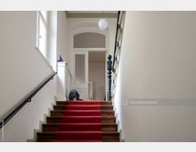 Treppenhaus in einem Altbau mit Holztreppe und rotem Teppichläufer, einer hellen Wandfarbe und Fenstern an der linken Seite. Am oberen Ende der Treppe befinden sich eine weiße Tür mit Glaseinsätzen und ein schwarzes Geländer mit verzierten Details.