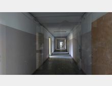 Langer, schmaler Flur in einem verlassenen Gebäude mit grauen Wänden, teils offenen Holztüren und einem Fliesenboden. Schwaches Tageslicht fällt durch die Türen und ein Fenster am Ende des Gangs.