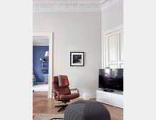 Bright living room with high ceilings, ornate white plaster crown molding, and pale walls, featuring a brown leather lounge chair and a large dark ottoman on a rug. A flat-screen TV sits on a low white media cabinet beside tall white paneled doors, with a wide doorway opening into a blue-walled room with shelving.