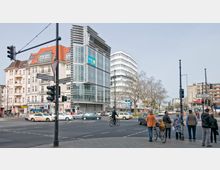 Straßenszene mit einer Kreuzung in einem urbanen Gebiet. Mehrstöckige Gebäude im Stil verschiedener Epochen, darunter ein moderner Glasbau, säumen die Straßen; Verkehrsschilder und Ampeln sind sichtbar, während Personen und Fahrzeuge die Kreuzung passieren.