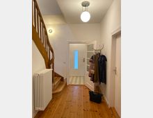 Ein Flur mit Holzdielenboden, einer hölzernen, geschwungenen Treppe auf der linken Seite und einer weißen Tür mit Glaseinsatz im Hintergrund. An der rechten Wand stehen eine Garderobe mit Jacken und Taschen sowie ein Heizkörper.