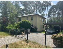 Zweistöckiges, hell verputztes Wohnhaus mit flachem Walmdach in einem grünen Garten, umgeben von hohen Bäumen. Vorn sind ein Metallzaun mit Einfahrtstor, gepflasterte Zufahrt und ein Carport neben dem Haus zu sehen.