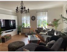 Gemütliches Wohnzimmer mit Holzboden, einer großen grauen Eckcouch, Zierkissen und einem Holztisch. An der Wand ein angebrachter Fernseher, darunter ein Lautsprecher; große Fenster mit weißen Vorhängen lassen Tageslicht hinein, umgeben von Zimmerpflanzen. Ein dekorativer Kronleuchter hängt von der Decke.